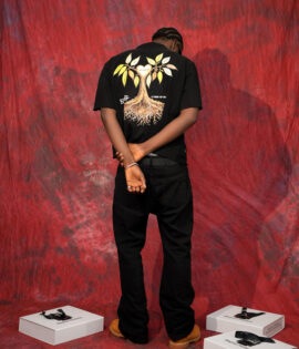 Model facing away in the BLAQFAM PREMIUM black shirt, displaying the tree back graphic against a red backdrop